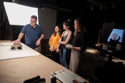 A teacher gestures toward a table in a lab with three students watching